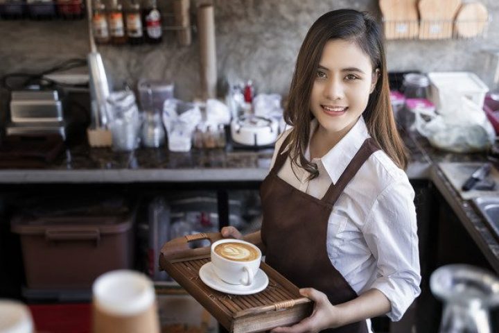 Young beautiful barista wearing brown apron holding hot coffee cup served to customer with smiling face at bar counter,Cafe restaurant service concept.waitress working